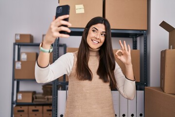 Canvas Print - Young brunette woman working at small business ecommerce taking selfie doing ok sign with fingers, smiling friendly gesturing excellent symbol