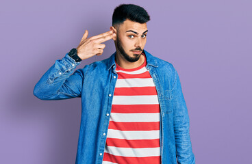 Young hispanic man with beard wearing casual denim jacket shooting and killing oneself pointing hand and fingers to head like gun, suicide gesture.