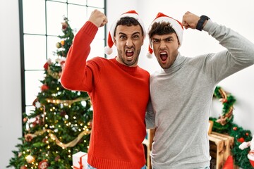 Poster - Young gay couple standing by christmas tree wearing hat angry and mad raising fist frustrated and furious while shouting with anger. rage and aggressive concept.