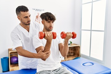 Canvas Print - Two hispanic men physiotherapist and patient having rehab session using dumbbells at clinic