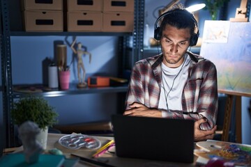 Poster - Young hispanic man sitting at art studio with laptop late at night skeptic and nervous, disapproving expression on face with crossed arms. negative person.