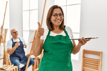 Sticker - Middle age hispanic woman at art studio smiling happy and positive, thumb up doing excellent and approval sign