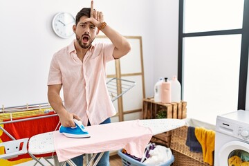 Poster - Young man with beard ironing clothes at home making fun of people with fingers on forehead doing loser gesture mocking and insulting.