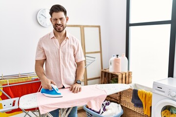 Sticker - Young man with beard ironing clothes at home winking looking at the camera with sexy expression, cheerful and happy face.