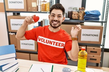 Poster - Young man with beard at donations stand holding red heart screaming proud, celebrating victory and success very excited with raised arm