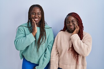 Poster - Two african woman standing over blue background touching mouth with hand with painful expression because of toothache or dental illness on teeth. dentist