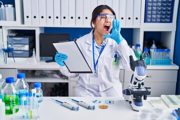 Canvas Print - Hispanic young woman working at scientist laboratory shouting and screaming loud to side with hand on mouth. communication concept.