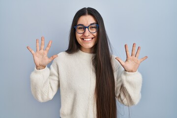 Poster - Young hispanic woman wearing casual sweater over blue background showing and pointing up with fingers number ten while smiling confident and happy.