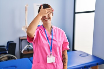 Canvas Print - Young brunette woman working at rehabilitation clinic smiling and laughing with hand on face covering eyes for surprise. blind concept.