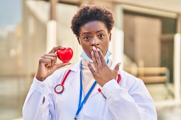 Poster - African american woman wearing doctor uniform holding heart covering mouth with hand, shocked and afraid for mistake. surprised expression