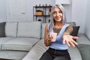 Canvas Print - Middle age grey-haired woman drinking coffee sitting on the sofa at home smiling cheerful offering palm hand giving assistance and acceptance.