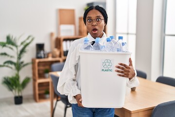 Wall Mural - African american woman working at the office holding plastic bottle for recycling afraid and shocked with surprise and amazed expression, fear and excited face.