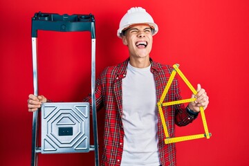 Wall Mural - Young hispanic man wearing handyman uniform holding construction stairs and house project angry and mad screaming frustrated and furious, shouting with anger looking up.