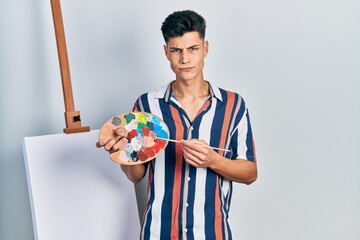 Canvas Print - Young hispanic man holding painter palette and paintbrush standing close to canvas skeptic and nervous, frowning upset because of problem. negative person.