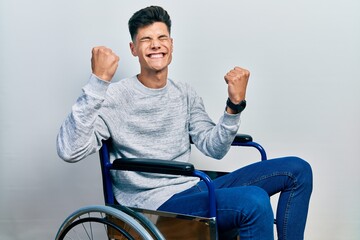 Wall Mural - Young hispanic man sitting on wheelchair very happy and excited doing winner gesture with arms raised, smiling and screaming for success. celebration concept.