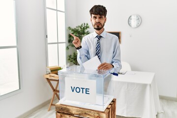 Wall Mural - Hispanic man with beard voting putting envelop in ballot box pointing aside worried and nervous with forefinger, concerned and surprised expression