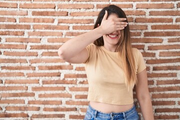 Poster - Young brunette woman standing over bricks wall smiling and laughing with hand on face covering eyes for surprise. blind concept.