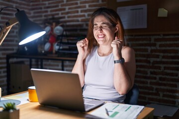 Poster - brunette woman working at the office at night excited for success with arms raised and eyes closed c