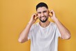 © Krakenimages.com - Young handsome man wearing casual t shirt over yellow background covering ears with fingers with annoyed expression for the noise of loud music. deaf concept.