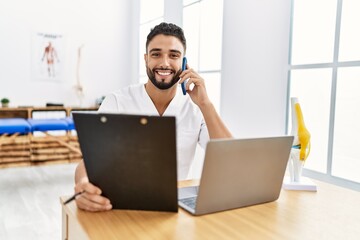 Sticker - Young arab man wearing physiotherapist uniform talking on the smartphone at clinic