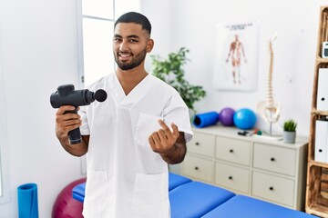 Poster - Young indian physiotherapist holding therapy massage gun at wellness center beckoning come here gesture with hand inviting welcoming happy and smiling