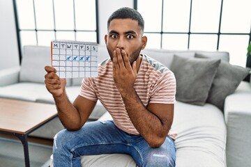 Sticker - Young indian man holding travel calendar covering mouth with hand, shocked and afraid for mistake. surprised expression