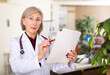 Portrait of experienced elderly female doctor standing in medical office with papers in hands, focused on studying clinical diagnosis of patient