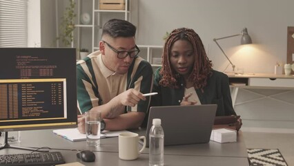Medium slowmo of multiethnic young couple of programmers sitting by laptop at office desk discussing and developing new software