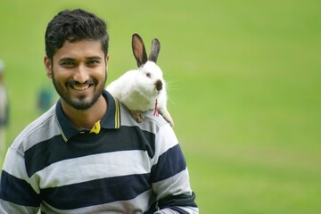 A handsome Indian Asian man, boy, male along with rabbit on shoulder showing animal, pet love. Nature, domestic, creature, friendly, happy, playful, smiling, background, zoo, garden, zoophilist, model