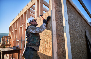 Wall Mural - Carpenter hammering nail into OSB panel on the wall of future cottage. Man worker building wooden frame house. Carpentry and construction concept.