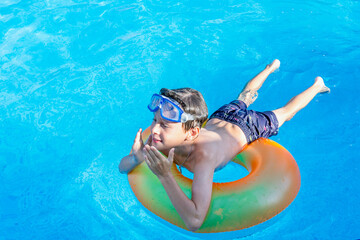 A joyful white Eastern European 9-year-old boy on a rainbow inflatable circle with swimming glasses enjoys life