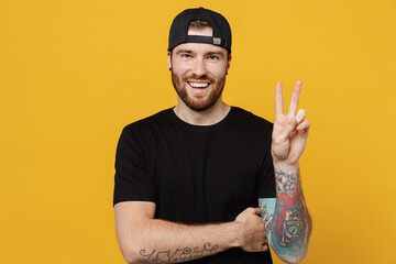 Wall Mural - Young smiling happy friendly positive bearded tattooed man 20s he wears casual black t-shirt cap showing victory sign isolated on plain yellow wall background studio portrait. Tattoo translate fun