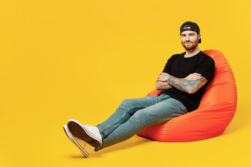 Wall Mural - Full body young bearded tattooed man 20s he wears casual black t-shirt cap sit in bag chair hold hands crossed folded isolated on plain yellow wall background studio portrait. Tattoo translate fun