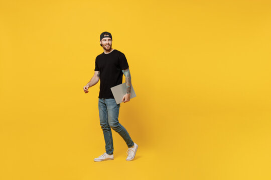 Full body young bearded tattooed man 20s he wear casual black t-shirt cap hold closed laptop pc computer look aside on workspace area isolated on plain yellow wall background People lifestyle concept