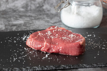 Rump beef steak, seasoned with sea salt with jar in background, on a slate chopping board. On a dark stone background