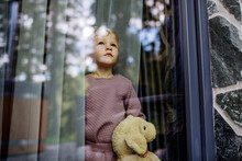Little Child Standing Alone With Teddy Bear Behind The Window, Photo Trough Glass.