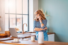 Happy Mature Woman Standing In The Kitchen At Home And Talking With Somebody On Her Smartphone