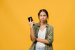 © tirachard - Thinking dreaming young Asia lady using phone with positive expression, dressed in casual cloth feeling happiness and stand isolated on yellow background. Happy adorable glad woman rejoices success.