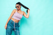 Leinwandbild Motiv Young beautiful smiling female in trendy summer jeans and top clothes. Carefree woman posing near blue wall in studio. Sexy positive model having fun indoors. Cheerful and happy. In sunglasses