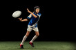 Dynamic portrait of school age boy, junior male rugby player practicing rugby football isolated on dark background with grass floooring.