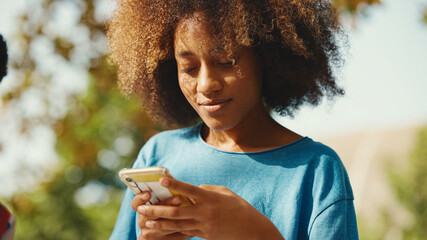 Wall Mural - Close-up of young smiling woman with curly hair sitting in park having picnic with friends on summer day outdoors. Girl uses cellphone, browses social networks, photo, video