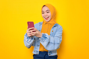 Wall Mural - Cheerful young Asian woman in jeans jacket looking at mobile phone screen, feels joyful reading good news isolated over yellow background