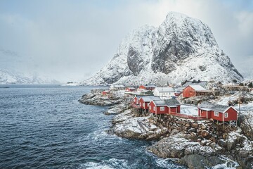 Sticker - View of red houses near water in Lofoten Islands, over background of snowy mountains in fog, Norway