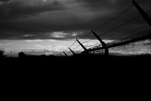 Black And White Shot Of Barbed Wire On Dark Fence Under Cloudy Sky. Monochrome Silhouette Photo