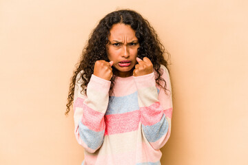 Wall Mural - Young hispanic woman isolated on beige background showing fist to camera, aggressive facial expression.