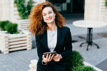 Young lady with curly hair, red painted lips and shining eyes, wearing black suit and shirt, sitting at outdoor cafe with tablet, smiling pleasantly into camera. Attractive female at terrace