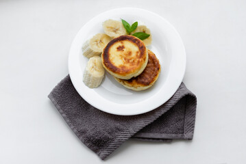 Two cottage cheese pancakes with sliced banana in a white plate. Traditional breakfast or lunch. Traditional Ukrainian breakfast.