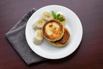 Two cottage cheese pancakes with sliced banana in a white plate. Traditional breakfast or lunch. Traditional Ukrainian breakfast.