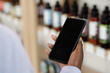 © pressmaster - Hand of young black woman holding smartphone while standing in front of display with variety of beautycare products in cosmetic shop