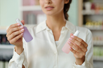 Wall Mural - Hands of young shopper holding two plastic bottles with liquid nailcare products in cosmetic shop while deciding which one to choose